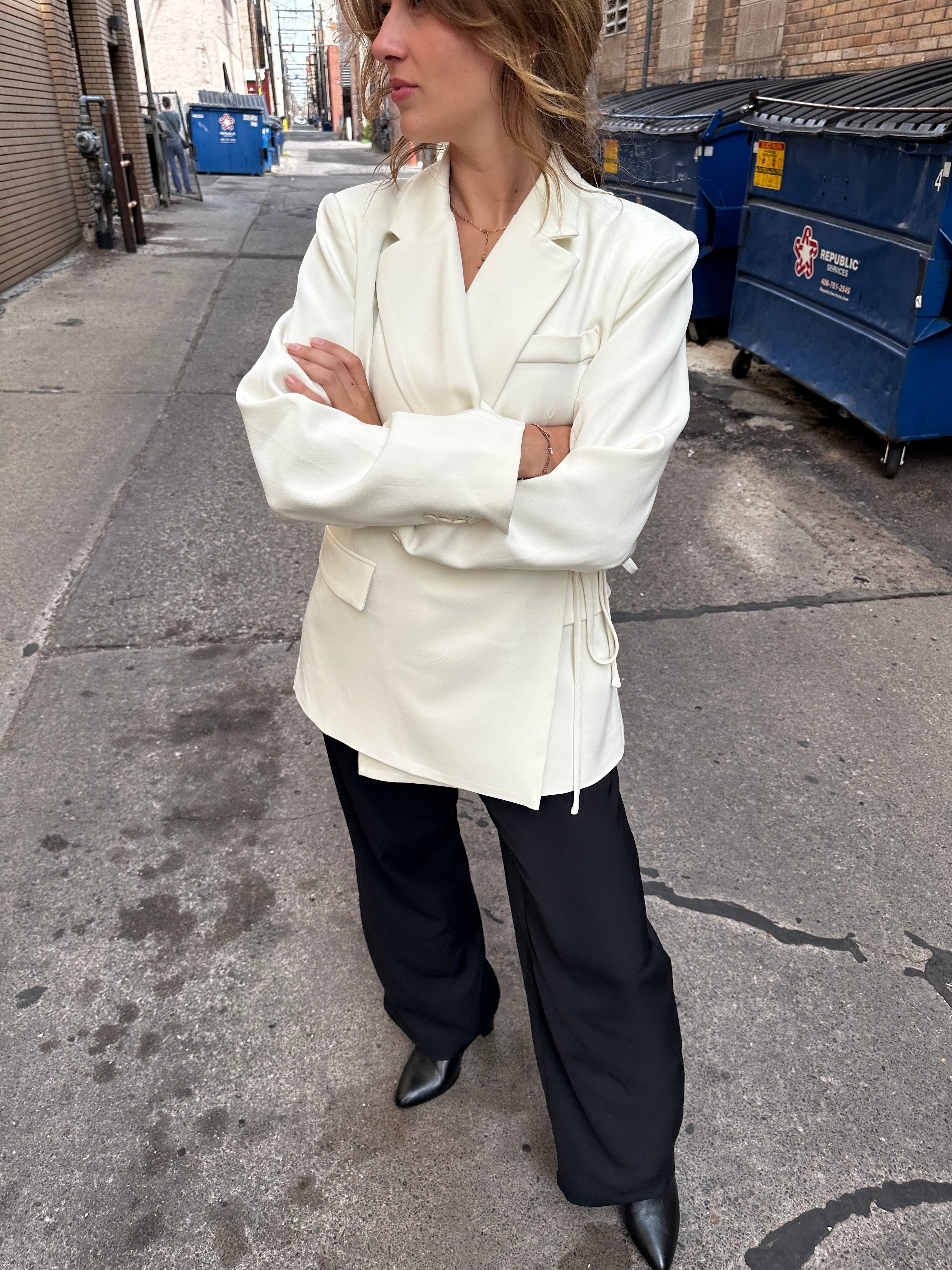 A woman in a cream blazer and black trousers with her arms crossed standing on the sidewalk.