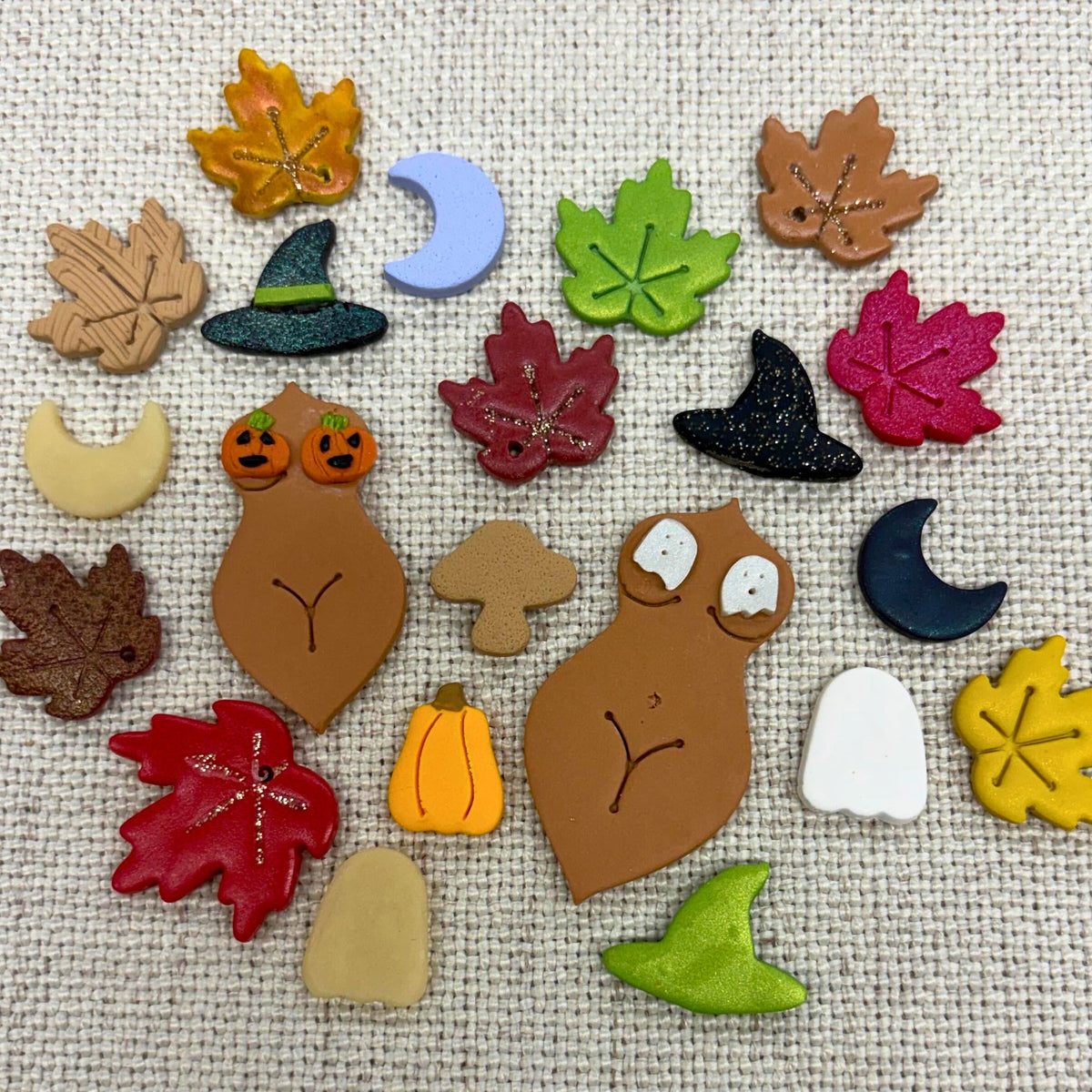 A collection of small autumn-themed polymer clay charms for a Great Falls, MT, fall-themed craft night. The charms are on a beige fabric background.