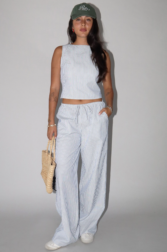 Woman wearing a light blue sleeveless top and wide-leg pants with a green cap, standing against a plain background.