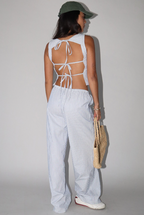 Person wearing a white open-back top and light blue striped pants with a green cap, standing against a plain background.