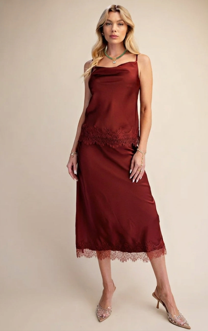 A woman models a sleeveless burgundy midi slip dress with delicate lace trim at the waist and hem, paired with sparkly pointed-toe heels against a solid beige studio background.
