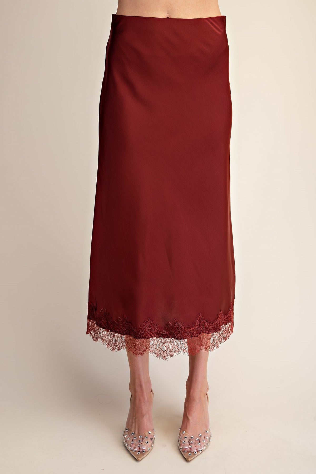 A dark red satin midi skirt with a delicate lace hem, shown on a model wearing clear embellished high heels against a plain beige background.