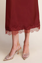 A close-up of a person's legs wearing a burgundy midi skirt with heels set against a plain beige background.