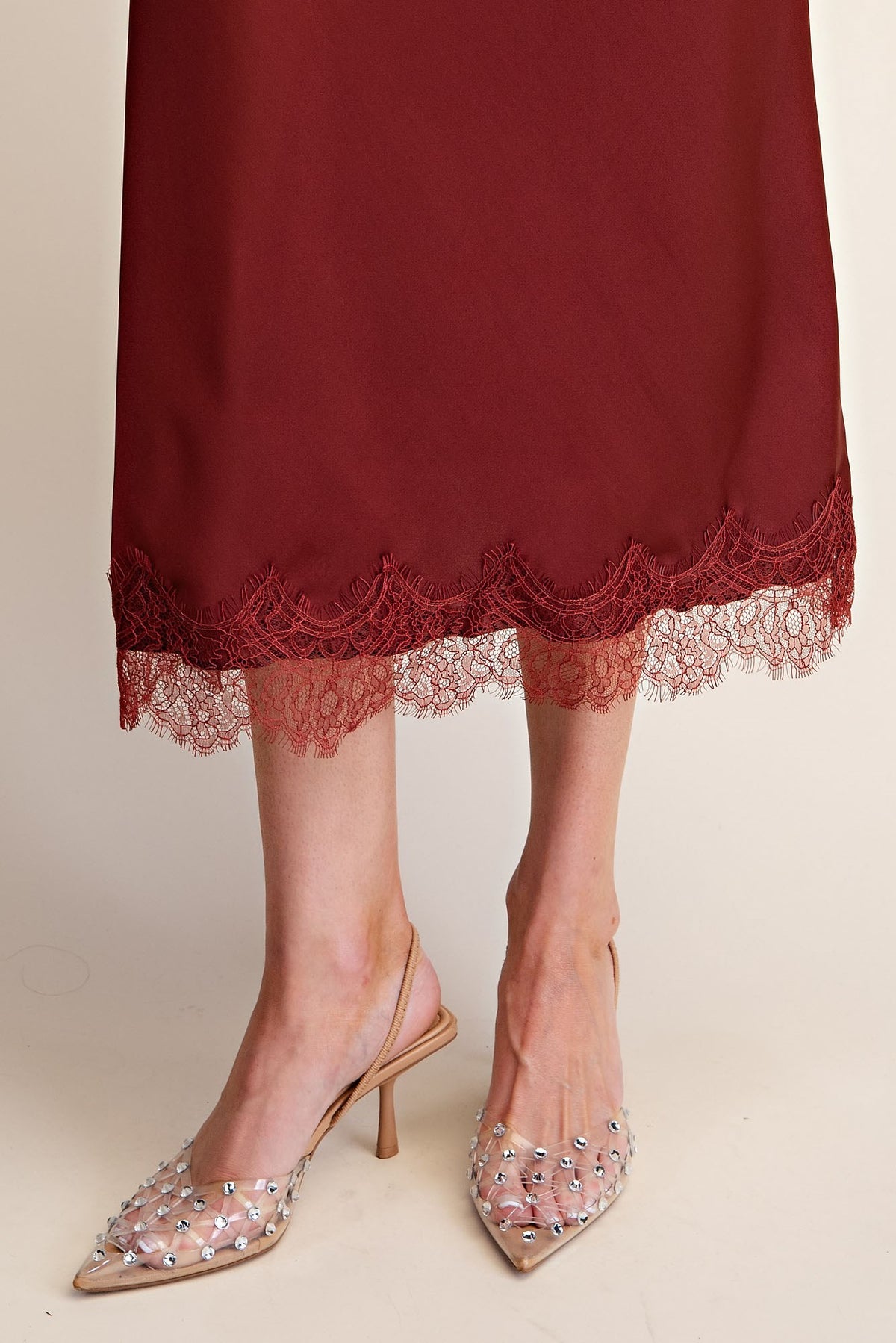 A close-up of a person's legs wearing a burgundy midi skirt with heels set against a plain beige background.