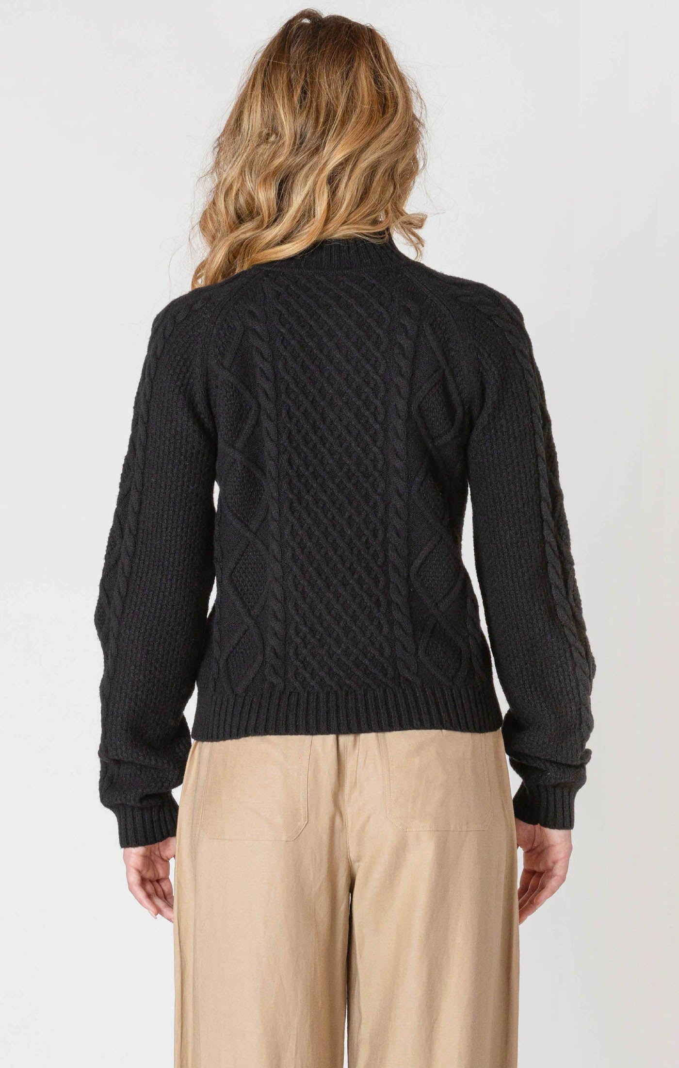 Person wearing a black cable knit sweater and beige pants on a white background