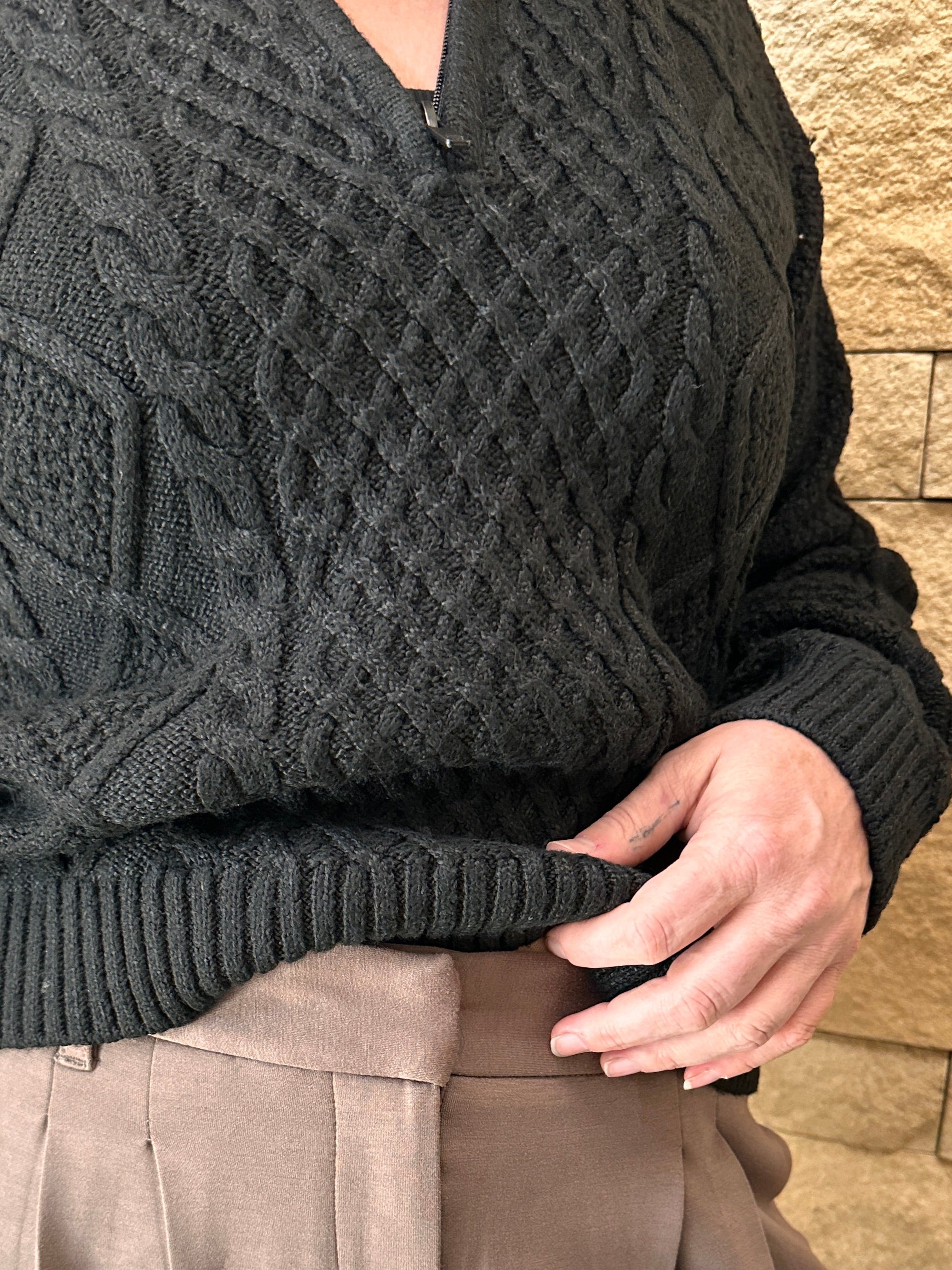 Person wearing a black textured sweater and beige pants with a stone wall background