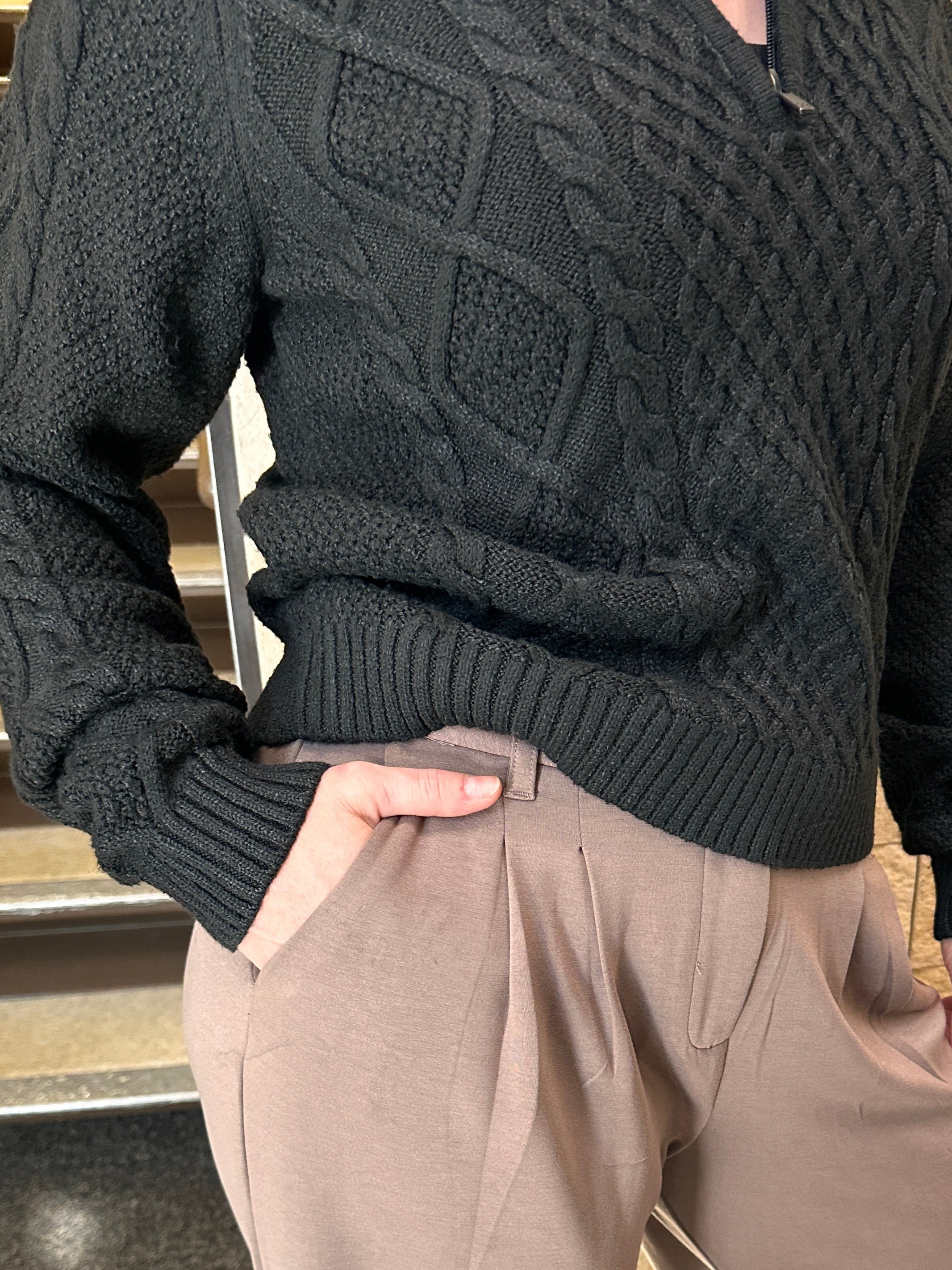 Person wearing a black cable knit sweater and beige pants on a wooden floor.