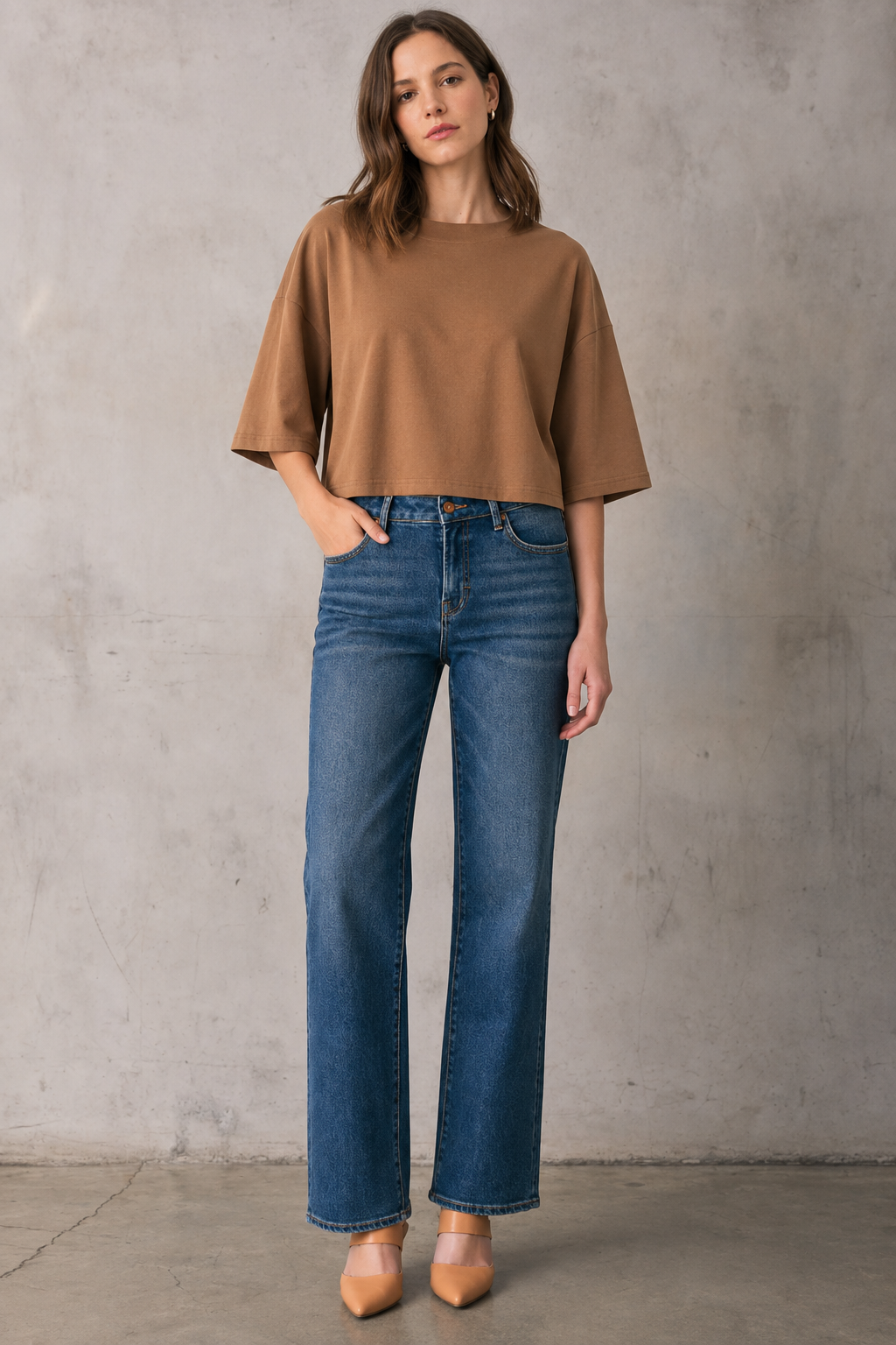 A woman stands against a textured gray concrete wall wearing a brown short-sleeve dropped shoulder top, blue straight-leg jeans, and tan pointed-toe heels.