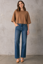 A woman stands against a textured gray concrete wall wearing a brown short-sleeve dropped shoulder top, blue straight-leg jeans, and tan pointed-toe heels.