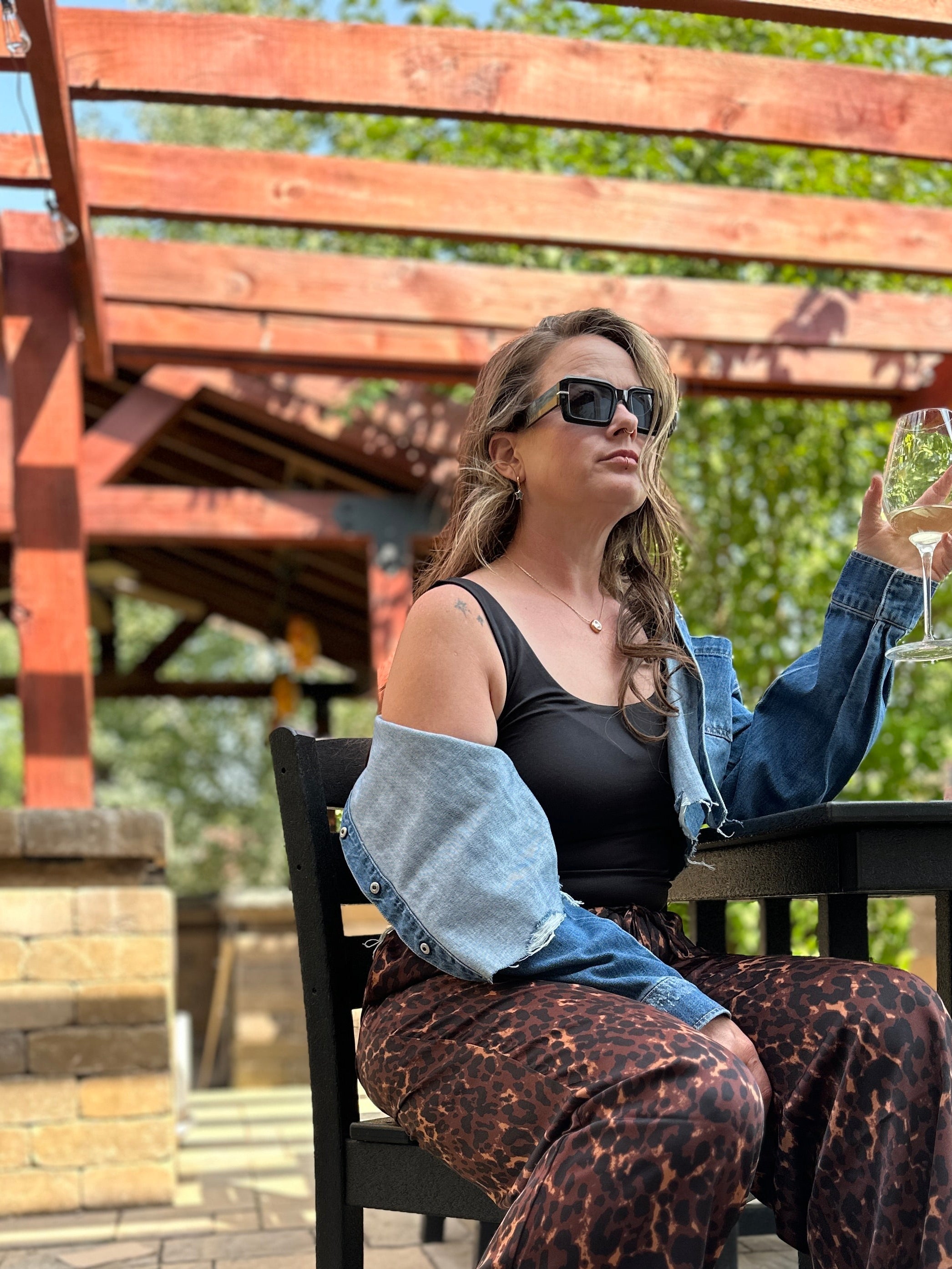 Woman sitting outdoors at a table holding a glass, wearing sunglasses and a denim jacket.