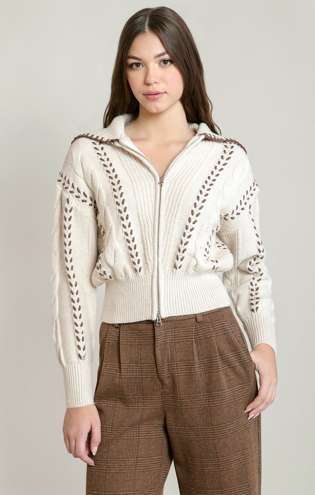 A woman modeling a cream-colored knit zip-up cable knit sweater with brown braided detailing on the sleeves and chest, paired with brown plaid trousers, against a light gray studio background.