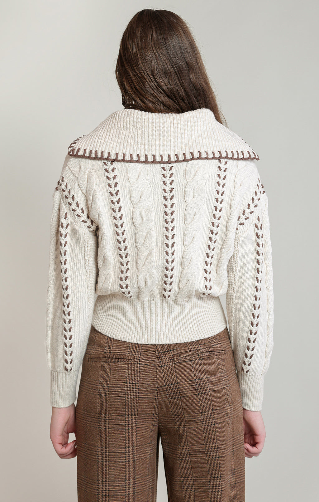 A woman shown from the back wearing a cream cable-knit cropped sweater with brown contrast stitching and a wide ribbed collar, paired with brown plaid trousers, against a light gray studio background.
