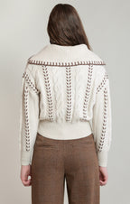 A woman shown from the back wearing a cream cable-knit cropped sweater with brown contrast stitching and a wide ribbed collar, paired with brown plaid trousers, against a light gray studio background.