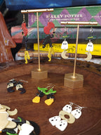 A collection of handmade polymer clay earrings displayed on gold-tone stands and arranged on a carved wooden tray, with Harry Potter books and ghost figurines in the background.