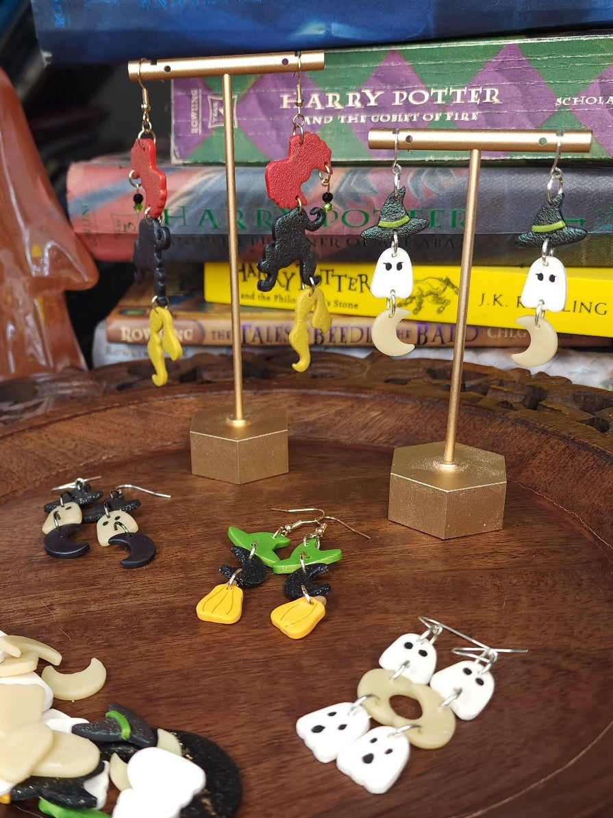 A collection of handmade polymer clay earrings displayed on gold-tone stands and arranged on a carved wooden tray, with Harry Potter books and ghost figurines in the background.