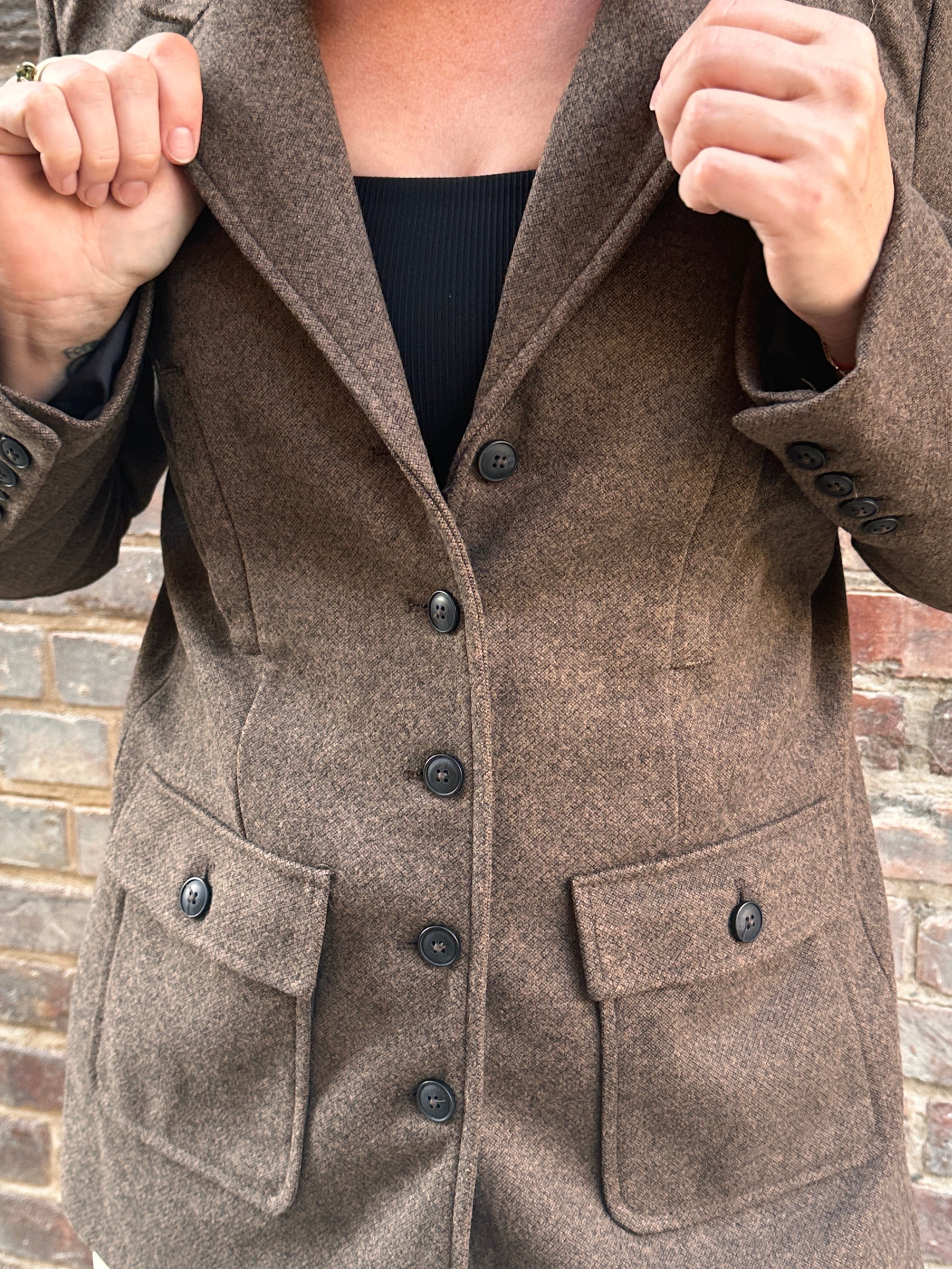 Close-up of a person wearing a brown tweed coat with button details on a brick wall background