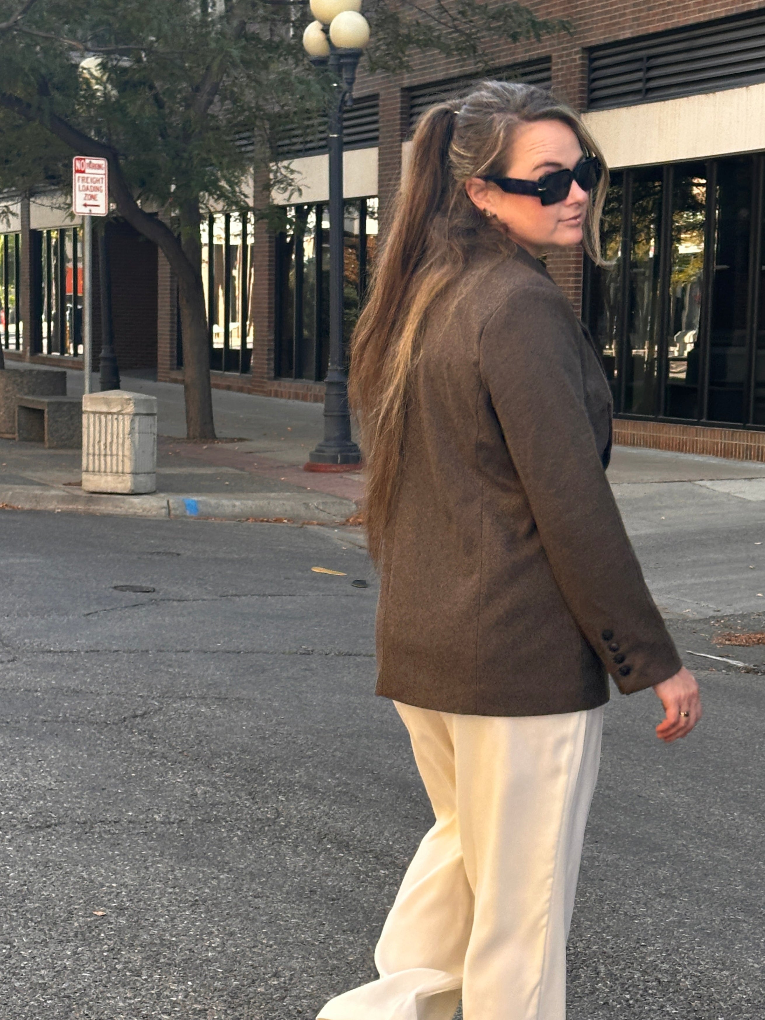 Woman walking down the street wearing sunglasses in a brown tweed blazer with cream pants.