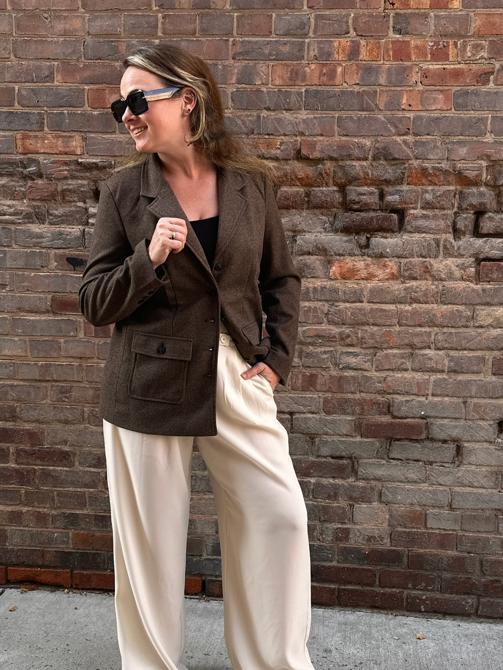 A woman standing in front of a brick wall wearing a brown tweed blazer with cream pants. Fall outfit