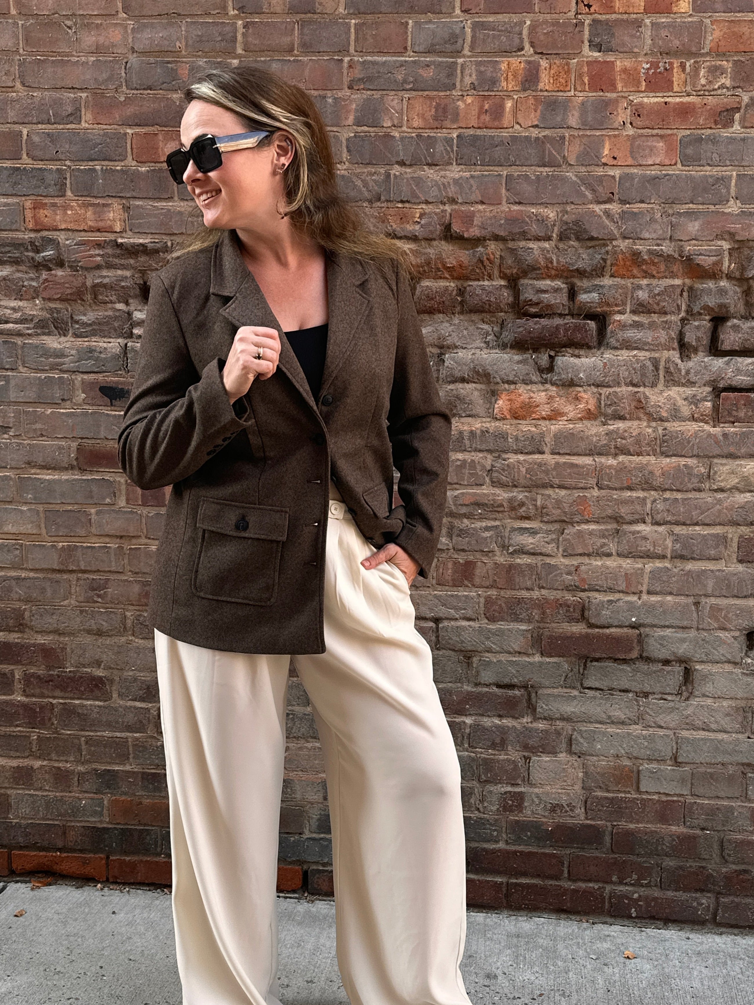 A woman standing in front of a brick wall wearing a brown tweed blazer with cream pants. Fall outfit