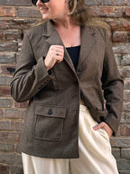 A woman wearing a brown tweed blazer with button details in front of a brick wall.