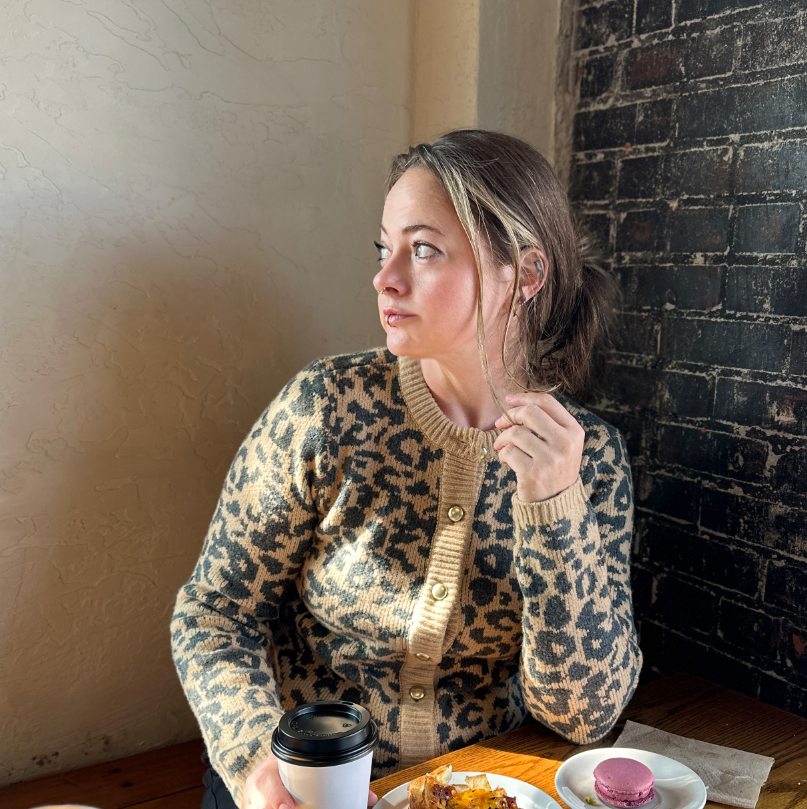 Woman in a leopard print cardigan sitting at a table with a coffee cup and dessert.