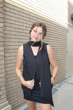 Woman wearing a black vest with scarf lapels standing on a sidewalk with a building in the background