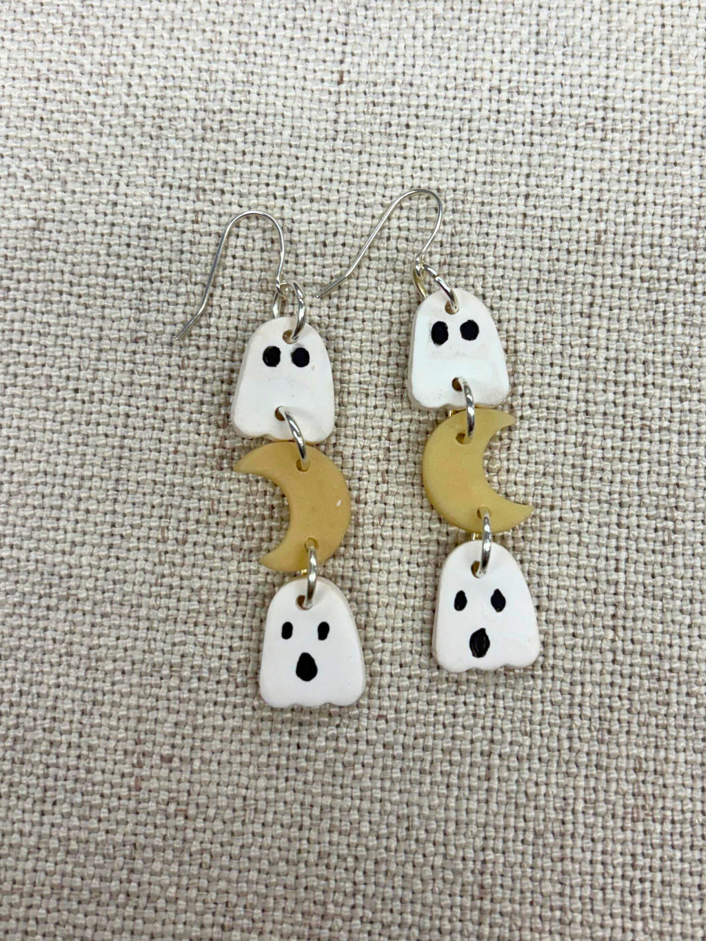 A pair of handmade dangle earrings featuring white ghost charms with black eyes and mouths, paired with small glow in the dark crescent moon charms, displayed on a textured beige fabric background.