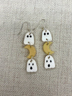 A pair of handmade dangle earrings featuring white ghost charms with black eyes and mouths, paired with small glow in the dark crescent moon charms, displayed on a textured beige fabric background.