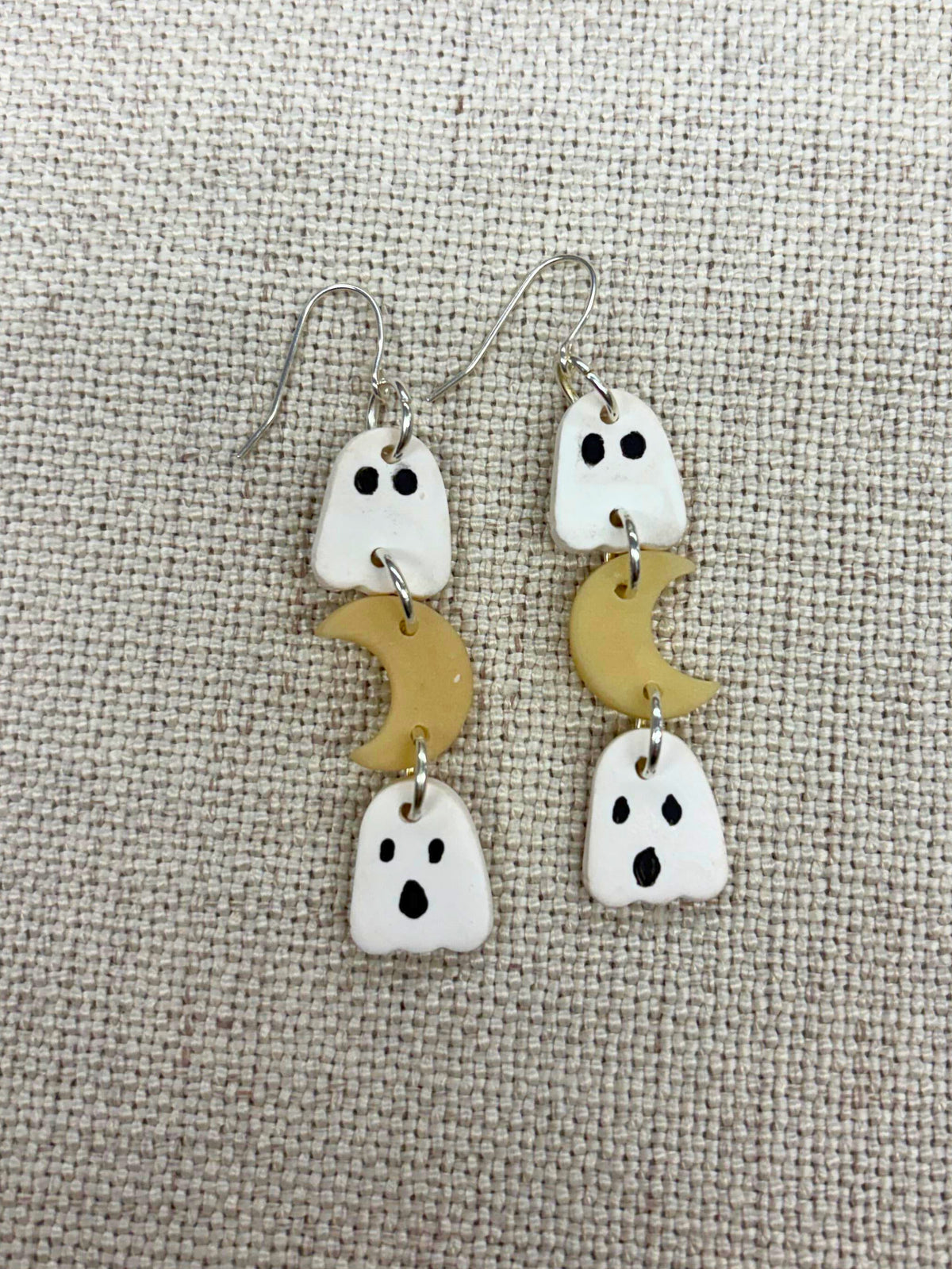 A pair of handmade dangle earrings featuring white ghost charms with black eyes and mouths, paired with small glow in the dark crescent moon charms, displayed on a textured beige fabric background.