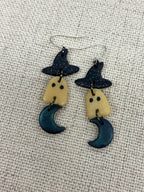 A pair of handmade dangle earrings featuring a witch's hat, a glow-in-the-dark ghost with black eyes, and a dark teal crescent moon, displayed on a textured beige fabric background.