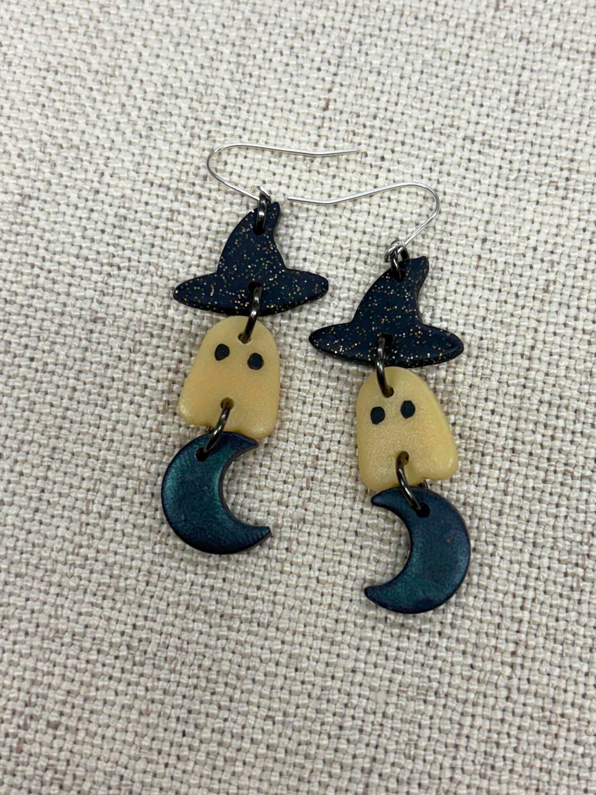 A pair of handmade dangle earrings featuring a witch's hat, a glow-in-the-dark ghost with black eyes, and a dark teal crescent moon, displayed on a textured beige fabric background.