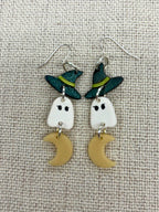 A pair of Halloween-themed dangle earrings featuring white ghost charms wearing green witch hats, with small glow-in-the-dark crescent moon charms below, displayed on a textured beige fabric background.