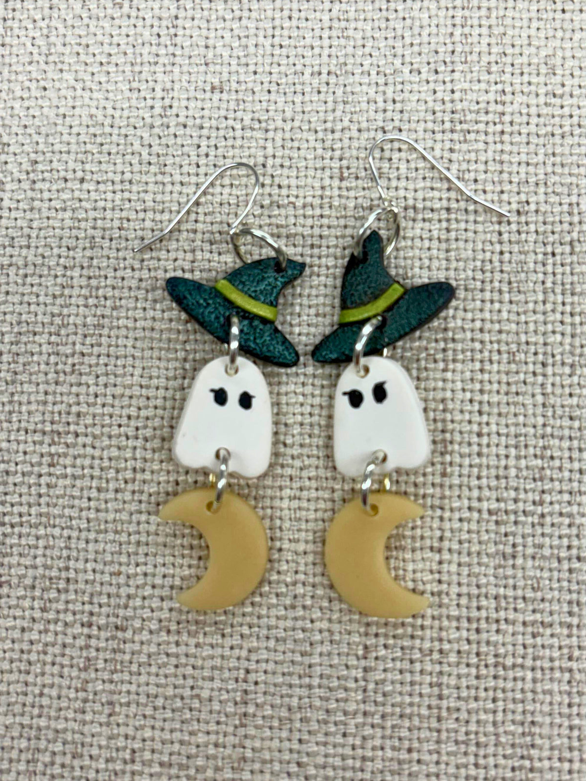 A pair of Halloween-themed dangle earrings featuring white ghost charms wearing green witch hats, with small glow-in-the-dark crescent moon charms below, displayed on a textured beige fabric background.