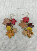 A pair of dangling earrings with autumn-themed charms, including red, brown, yellow, and tan maple leaves and acorns, hanging from gold-tone hooks on a textured off-white fabric background.