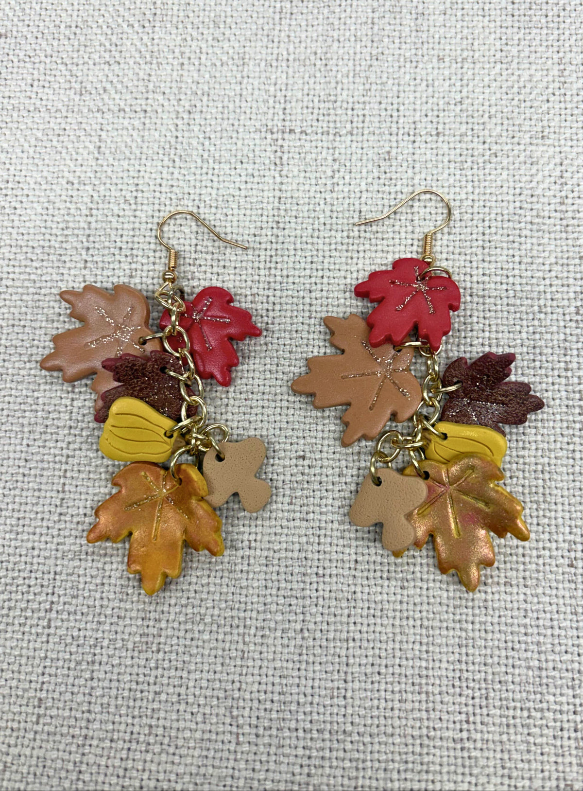A pair of dangling earrings with autumn-themed charms, including red, brown, yellow, and tan maple leaves and acorns, hanging from gold-tone hooks on a textured off-white fabric background.