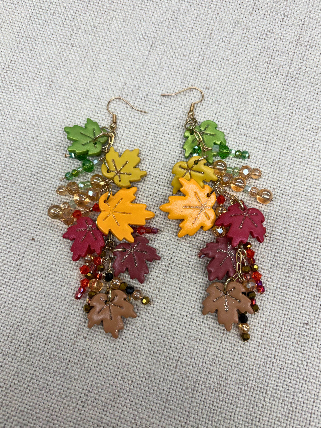 A pair of dangling earrings with colorful autumn leaf charms in green, yellow, burgundy, and brown, accented with small beads and gold-tone hooks, displayed on a textured beige fabric background.