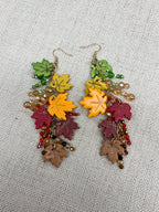 A pair of dangling earrings with colorful autumn leaf charms in green, yellow, burgundy, and brown, accented with small beads and gold-tone hooks, displayed on a textured beige fabric background.