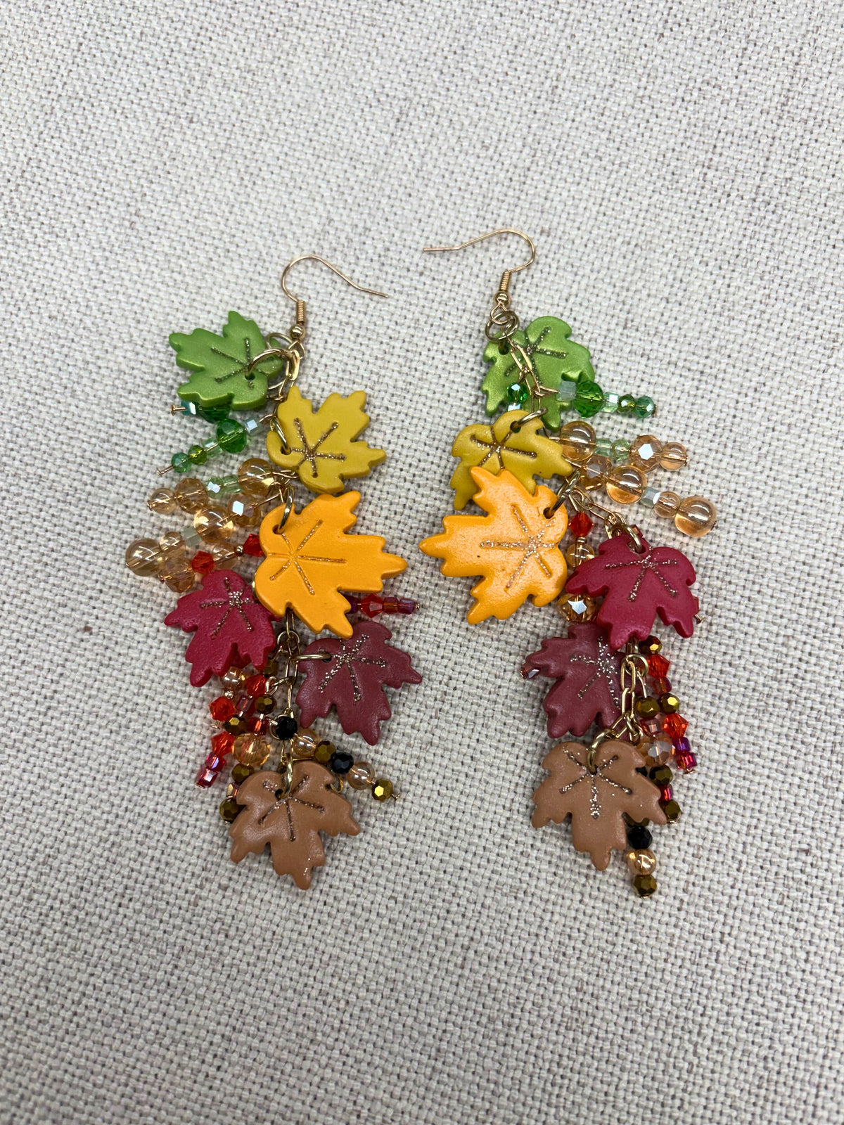 A pair of dangling earrings with colorful autumn leaf charms in green, yellow, burgundy, and brown, accented with small beads and gold-tone hooks, displayed on a textured beige fabric background.