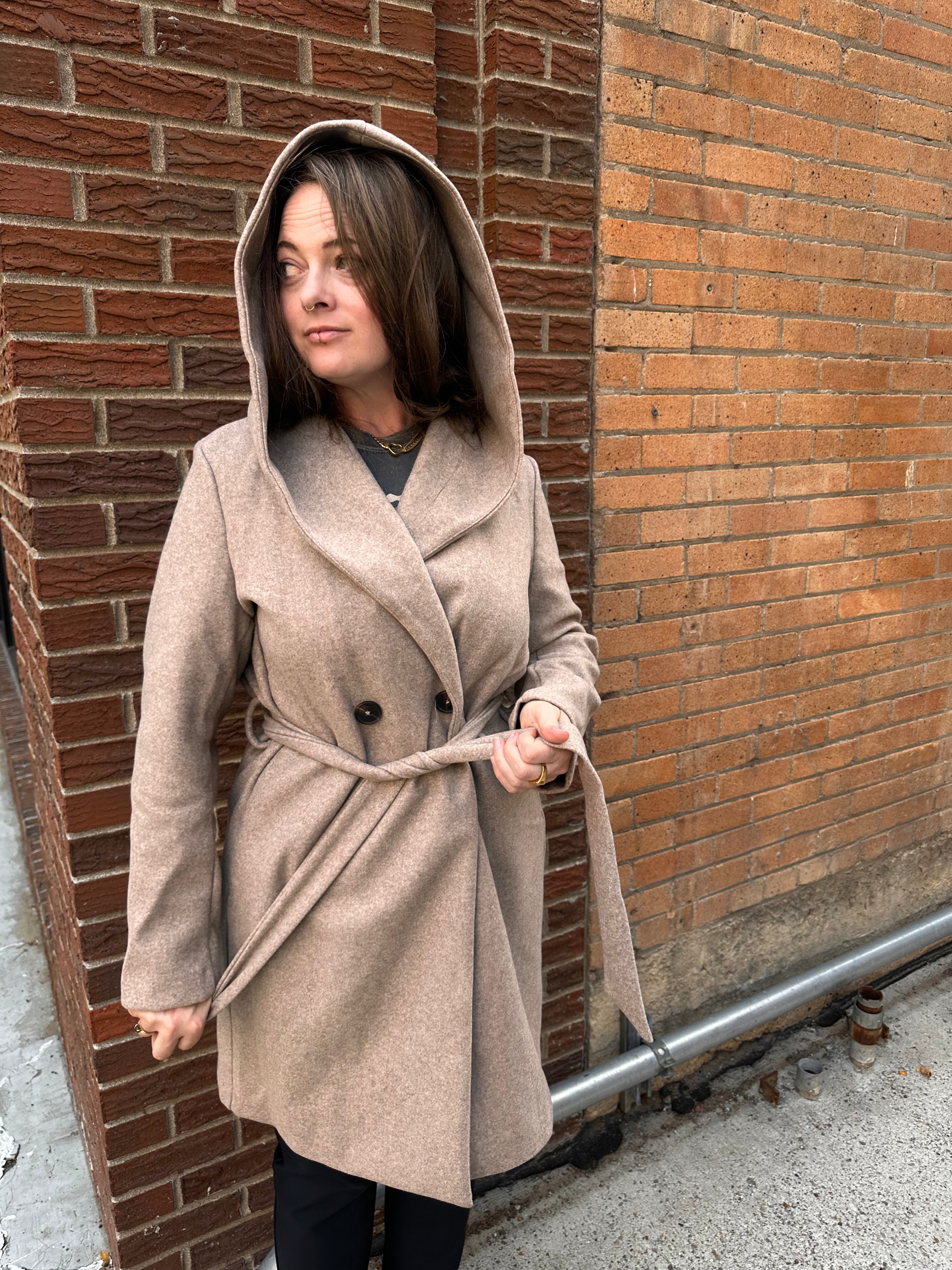 Woman wearing a beige coat with a hood against a brick wall.