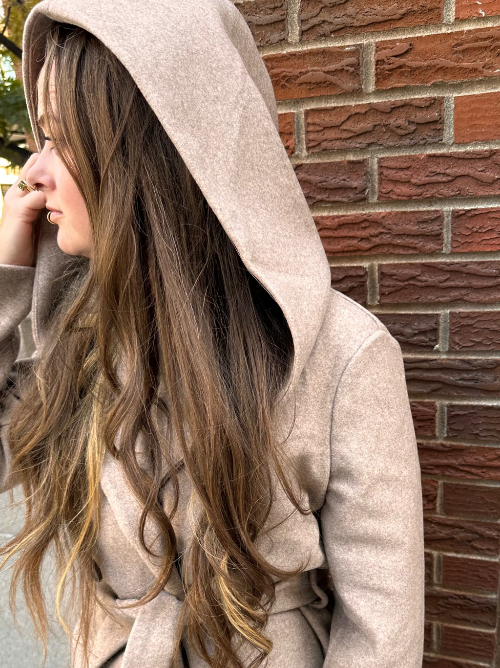 Person wearing a beige hooded coat standing against a brick wall.