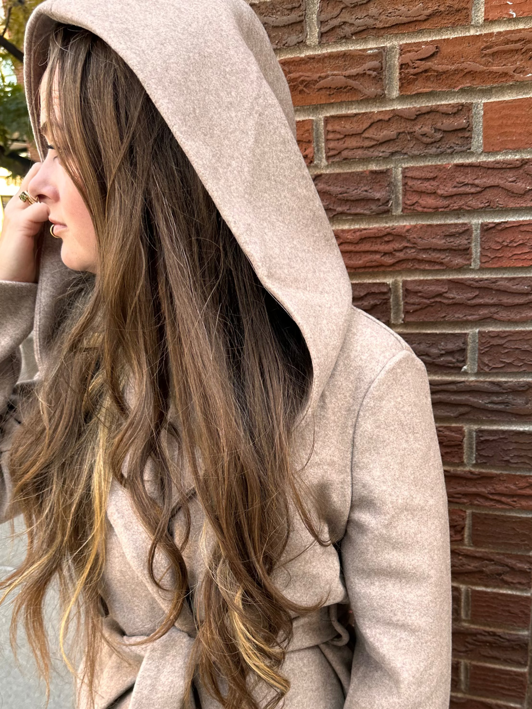 Person wearing a beige hooded coat standing against a brick wall.