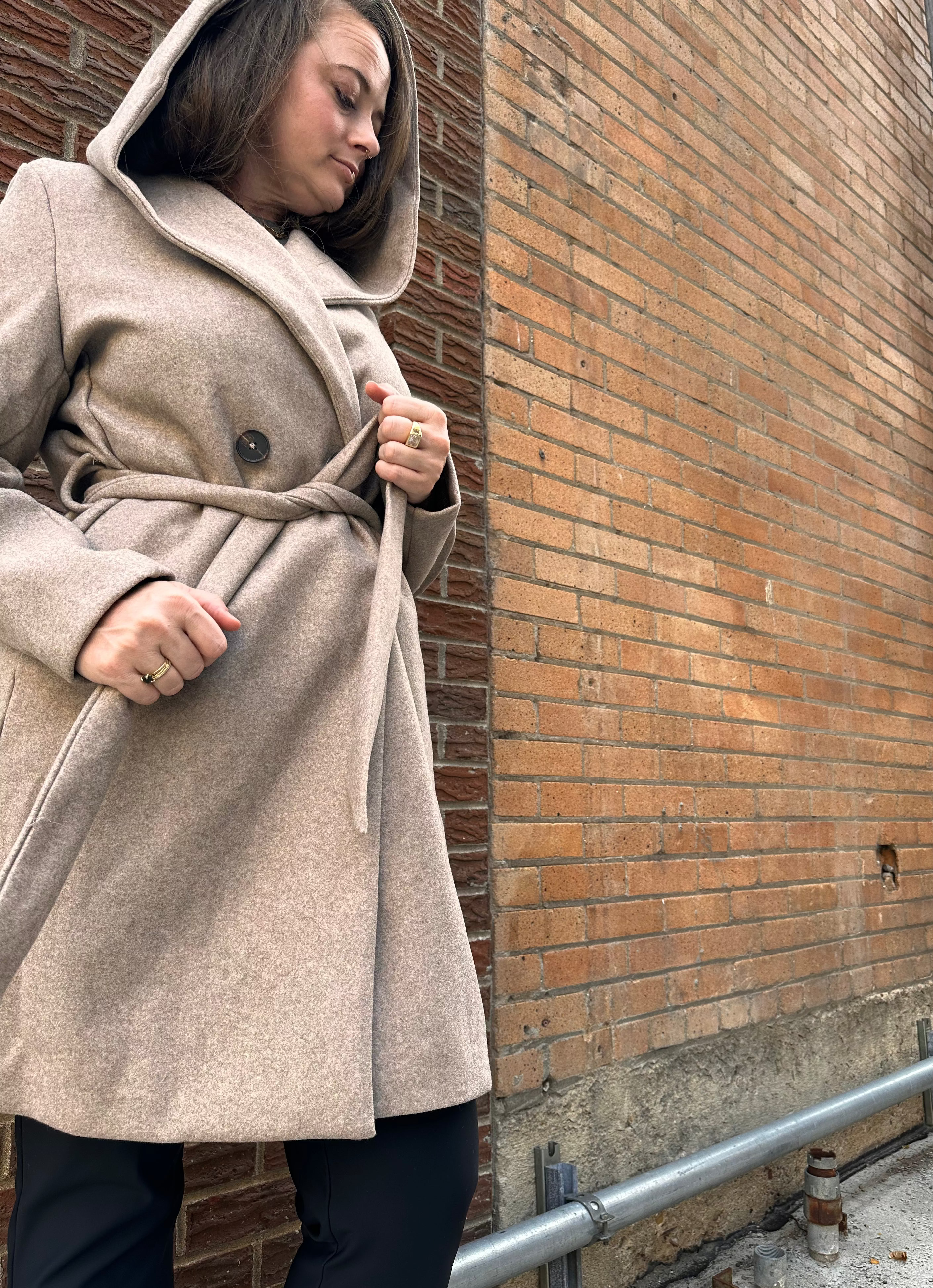 Woman in a beige coat standing against a brick wall.