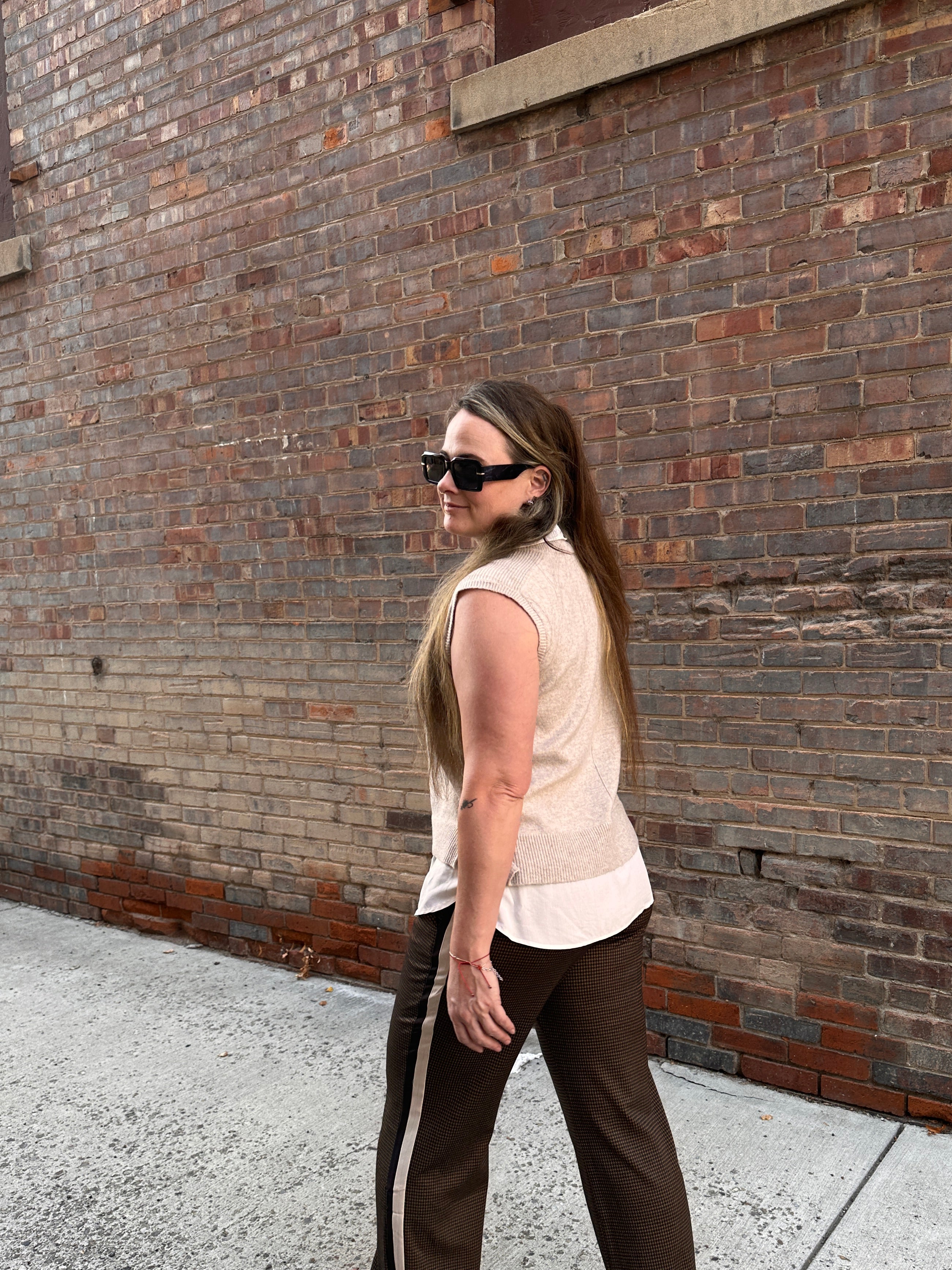 A woman wearing a sweater vest and pants with a stripe down the side walking on a sidewalk with a brick wall in the background