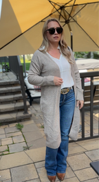 Woman wearing a long beige cardigan, white shirt, blue jeans, and sunglasses outdoors.