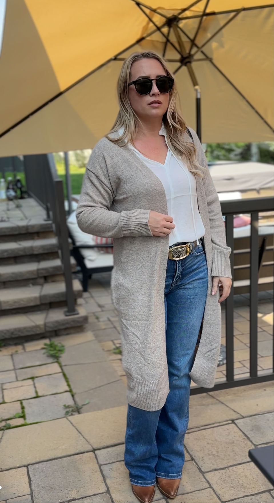 Woman wearing a long beige cardigan, white shirt, blue jeans, and sunglasses outdoors.