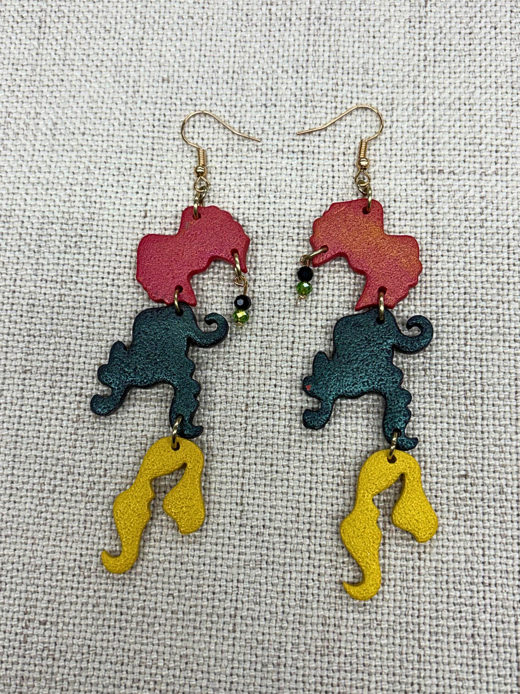 A pair of dangling earrings laid flat on a textured beige fabric background. Each earring has a gold-tone fishhook wire with Hocus Pocus themed custom charms.