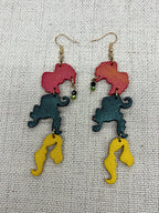 A pair of dangling earrings laid flat on a textured beige fabric background. Each earring has a gold-tone fishhook wire with Hocus Pocus themed custom charms.