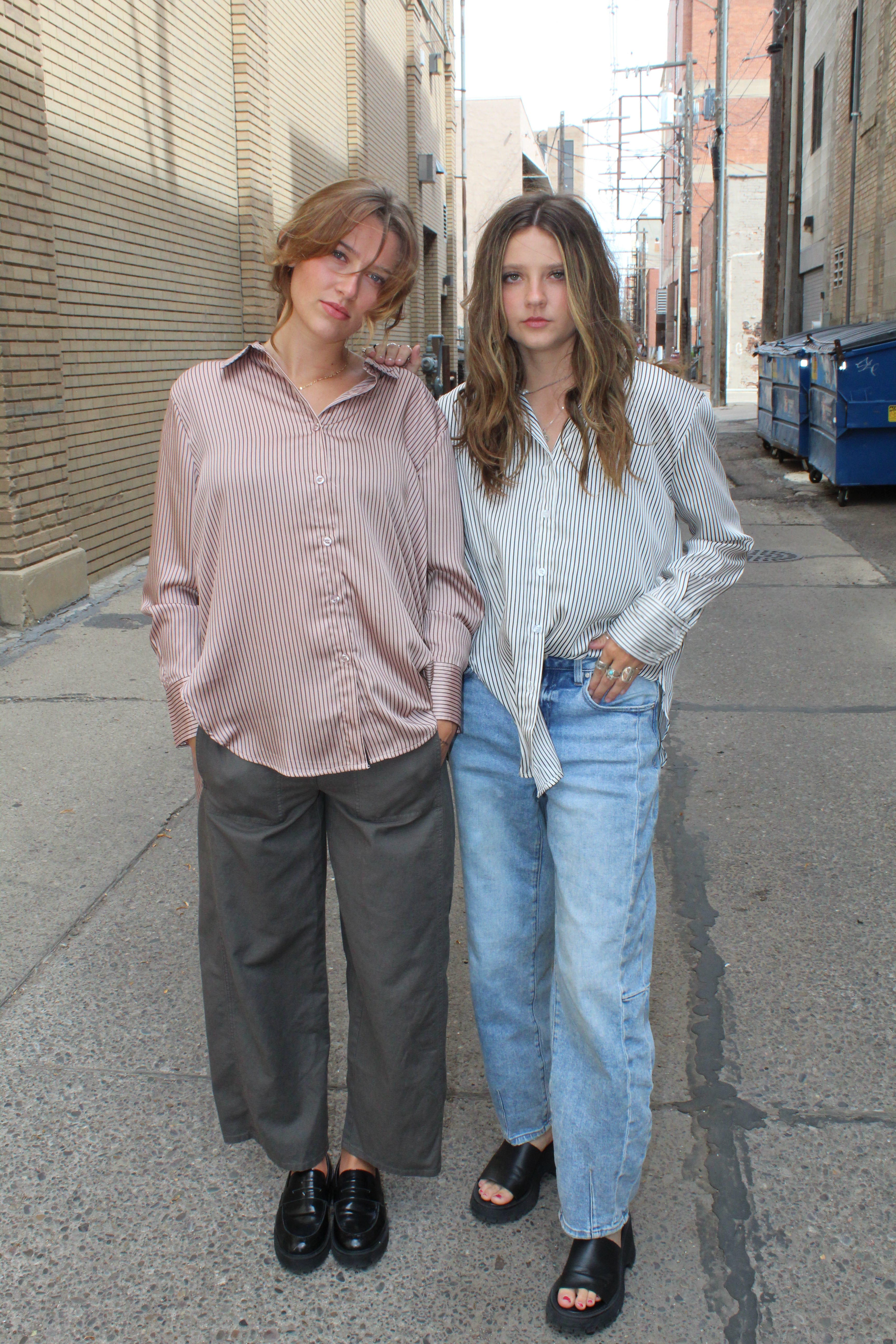 Two people standing on a sidewalk with a building in the background, one wearing a rose vertical striped button up and the other wearing a black and white vertical striped button up. 