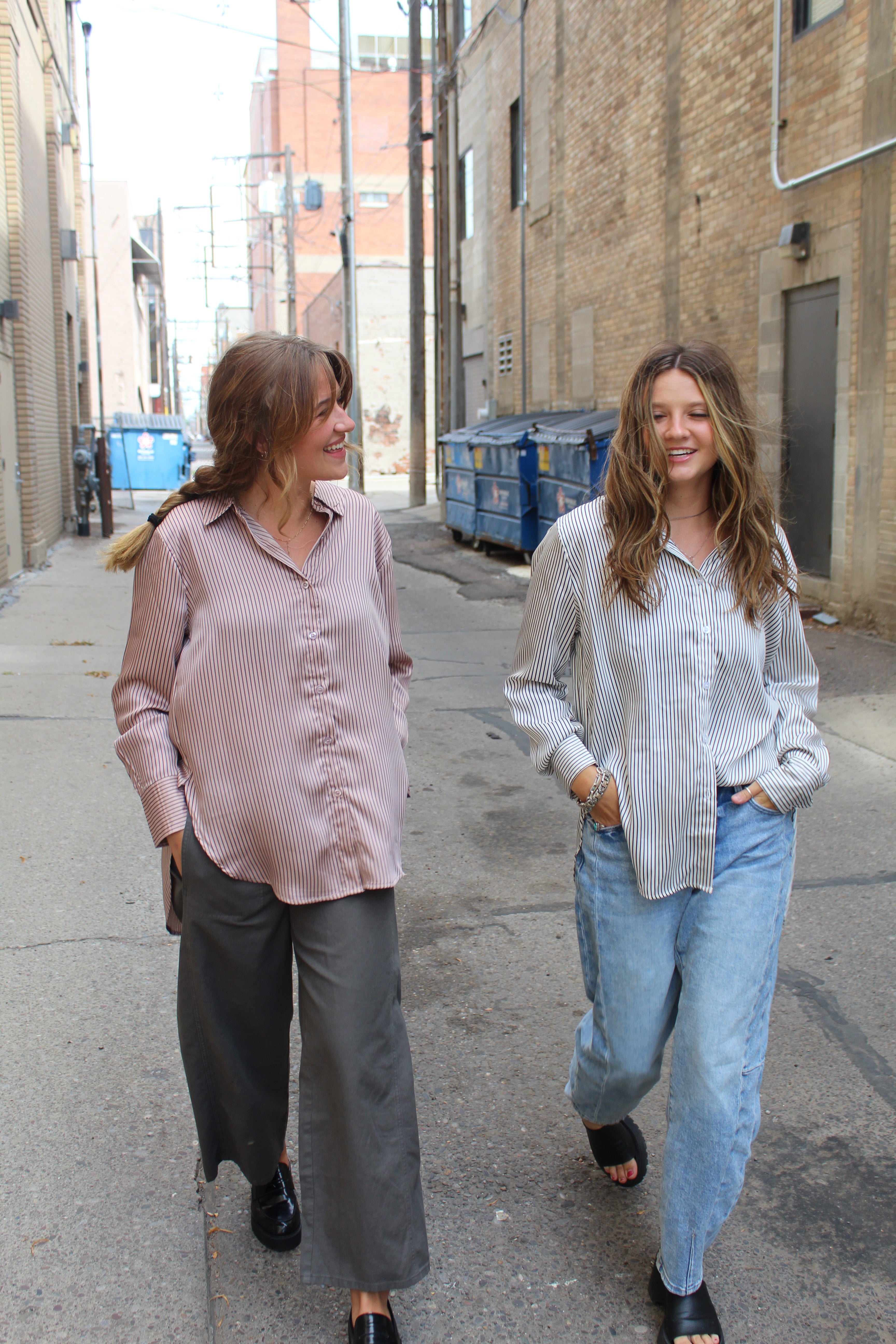 Two people standing on a sidewalk with a building in the background, one wearing a rose vertical striped button up and the other wearing a black and white vertical striped button up. 
