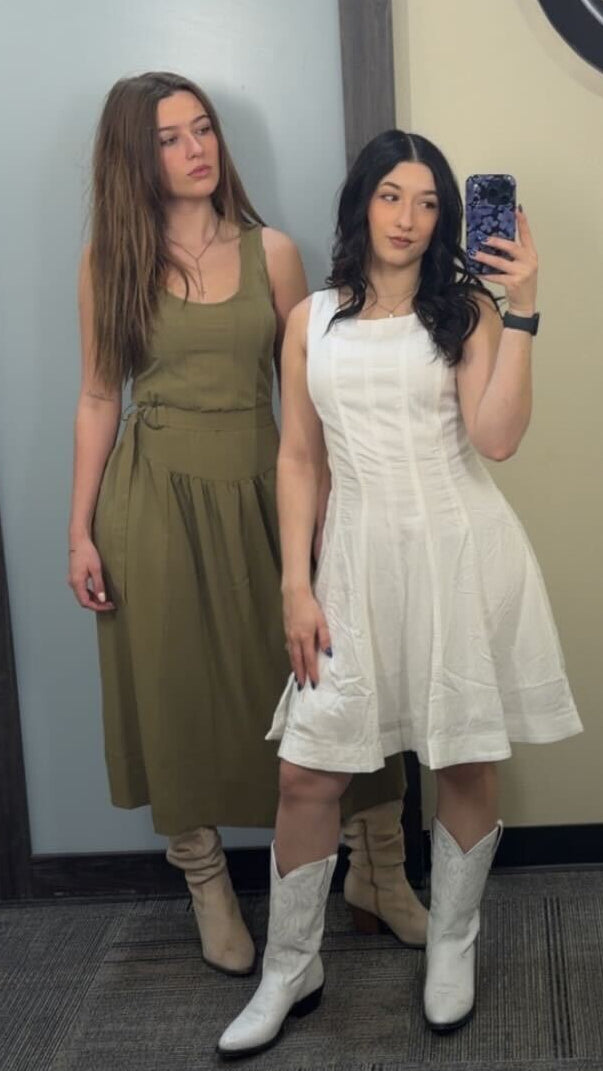 Two women standing in front of a mirror wearing dresses, with a floral-patterned tablecloth in the foreground.
